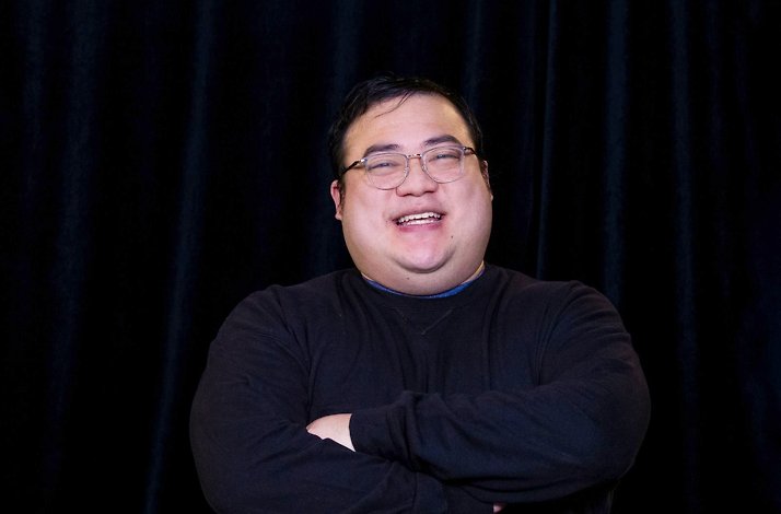 Scarra smiling with arms crossed, wearing glasses and a black turtleneck, posed against a dark draped backdrop.