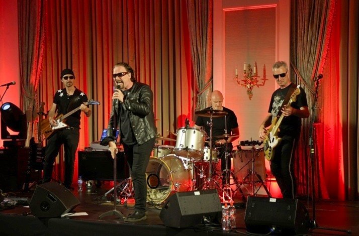 U2 band performs on stage with red lighting, guitars, drums, and a vocalist in focus.