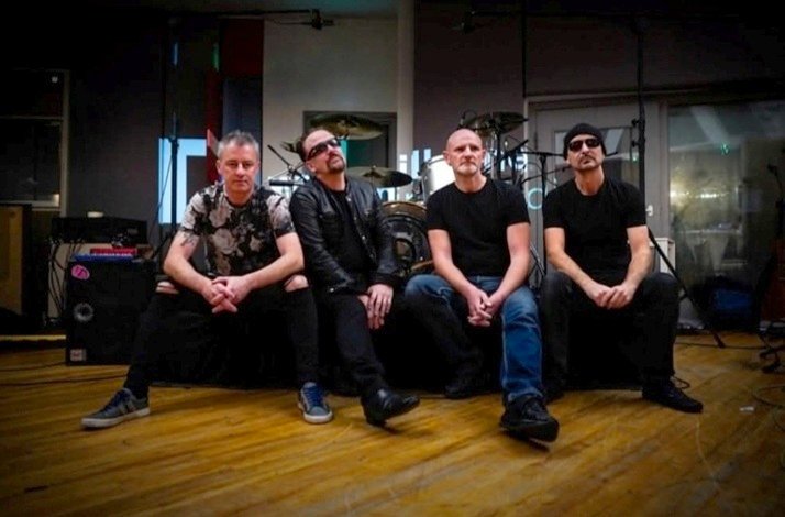 Band members are seated indoors with musical equipment and drums in the background.