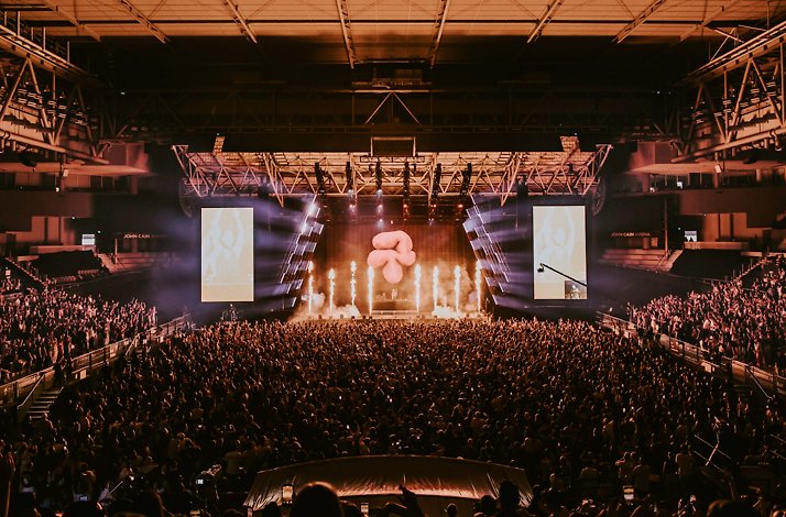 Packed crowd at John Cain Arena during AO Live Presents, with pyrotechnics, beam lighting, and a large inflatable prop illuminating the stage.