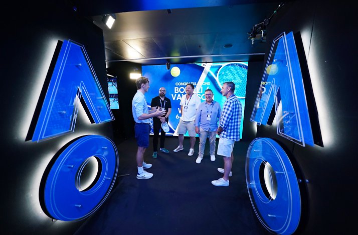 Five people standing and talking in a dimly lit room with a blue AO ign on the wall and a screen behind them.