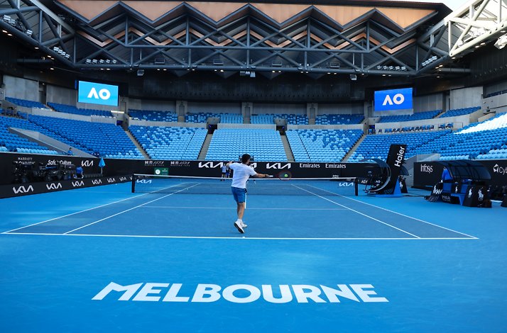 A tennis player on a blue court with a racket in hand, with empty stadium seats in the background.