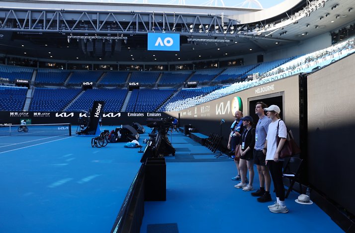 Four people standing by a doorway on the edge of a tennis court inside an empty stadium.