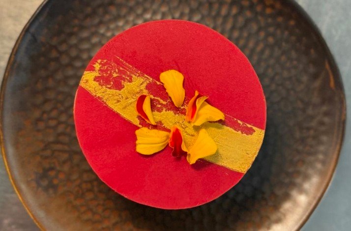 A vibrant dessert with a red and gold design, adorned with yellow flower petals on a textured black plate