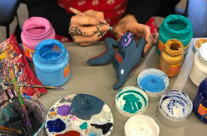 The artist is painting a small gray figurine with blue and pink dots.