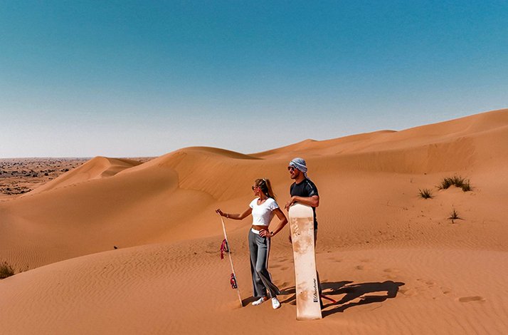 Two people holding sandboards, standing on sand dunes under a clear blue sky.