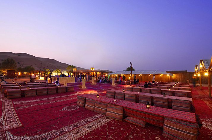 Desert camp setup with traditional seating, rugs, and ambient lighting at sunset.