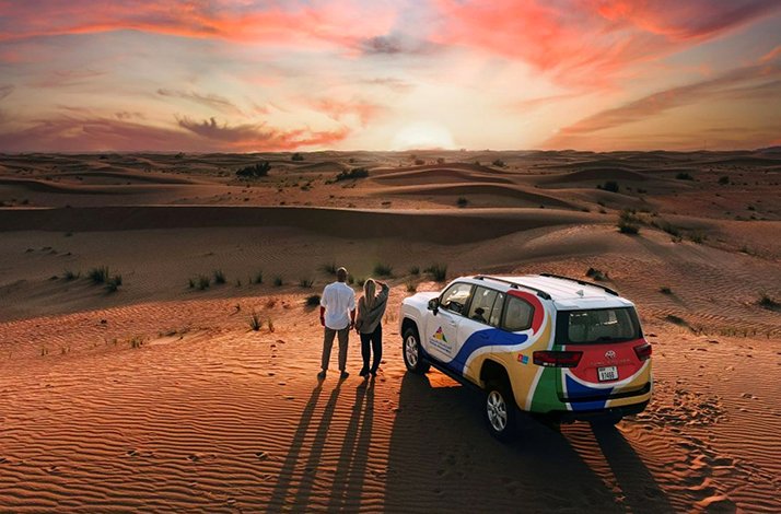 An SUV parked in the Dubai Desert Conservation Reserve as people admire the sunset over sand dunes.