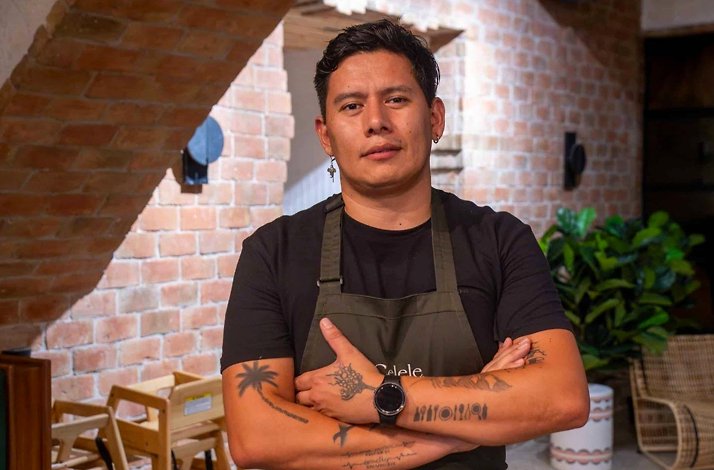 A chef in a Celele apron standing inside the restaurant's exposed-brick dining room with wooden furniture and plants, at Celele in Cartagena, Colombia