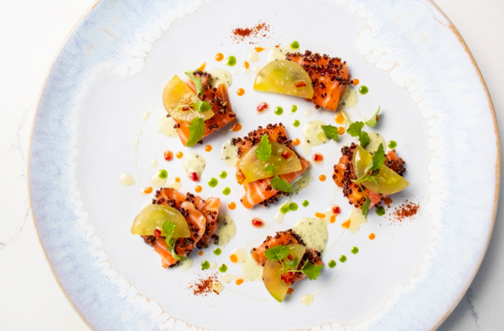 Salmon pieces crusted with black quinoa, green tomato wedges, herb cream, and micro mint on a pale blue plate.