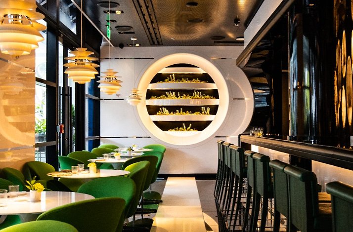 Le Jardinier Miami dining room with emerald green chairs, a lit circular herb wall feature, and a dark bar area.