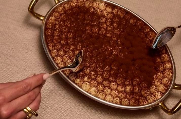 A large, oval-shaped serving dish filled with a creamy tiramisu dusted with cocoa powder.