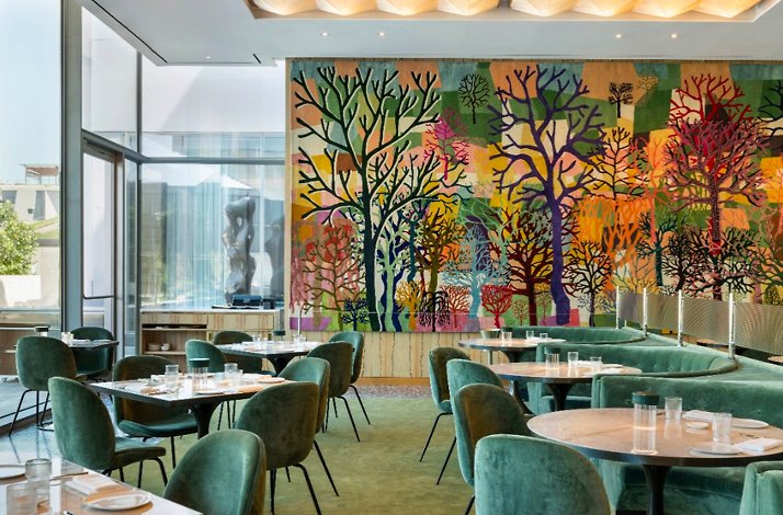 Sunlit dining room with green velvet seating and a colorful tree mural at Le Jardinier Houston.