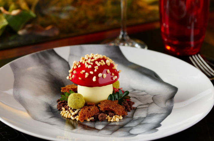 Artfully plated mushroom-shaped dessert with glossy red cap and crisp garnishes on a black-and-white patterned plate.