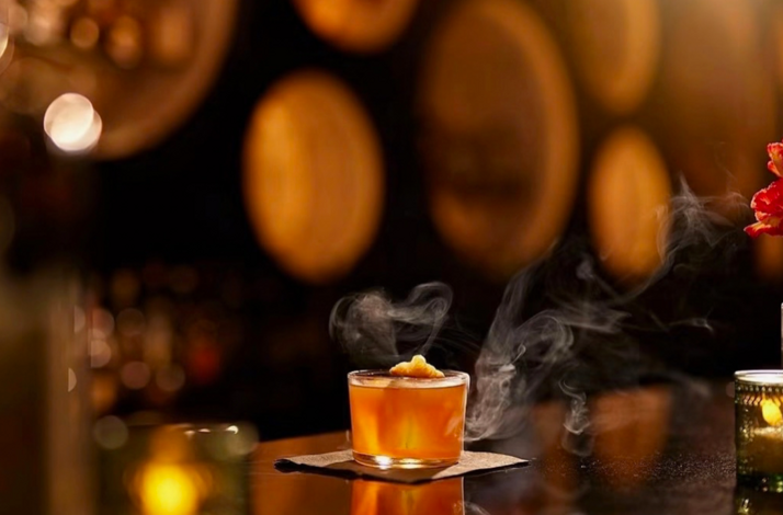 Smoked amber cocktail in a lowball glass on a bar top, with warm bokeh lights and floral accents in the background.