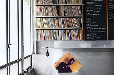Record shelf behind a metal counter; chalkboard menu at right and a vinyl sleeve leaning in front.